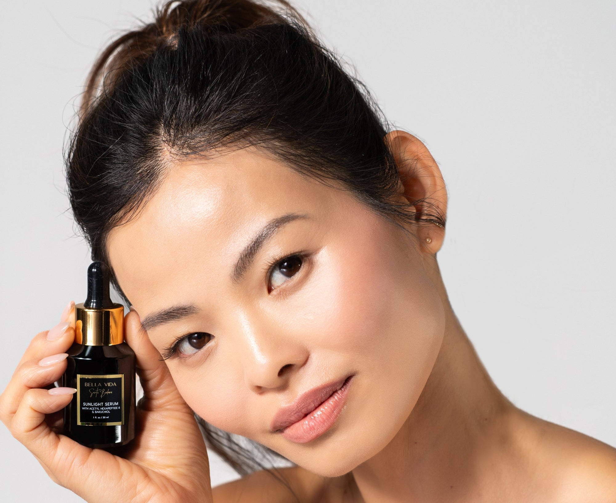 Woman holding a Black serum bottle with dropper labeled 'Bella Vida Santa Barbara Sunlight Serum with Acetyl Hexapeptide-8 and Bakuchiol . Sunlight Serum Botox Alternative without injections. Reduces wrinkles, brightens and smooths skin, hydrates, increases collagen