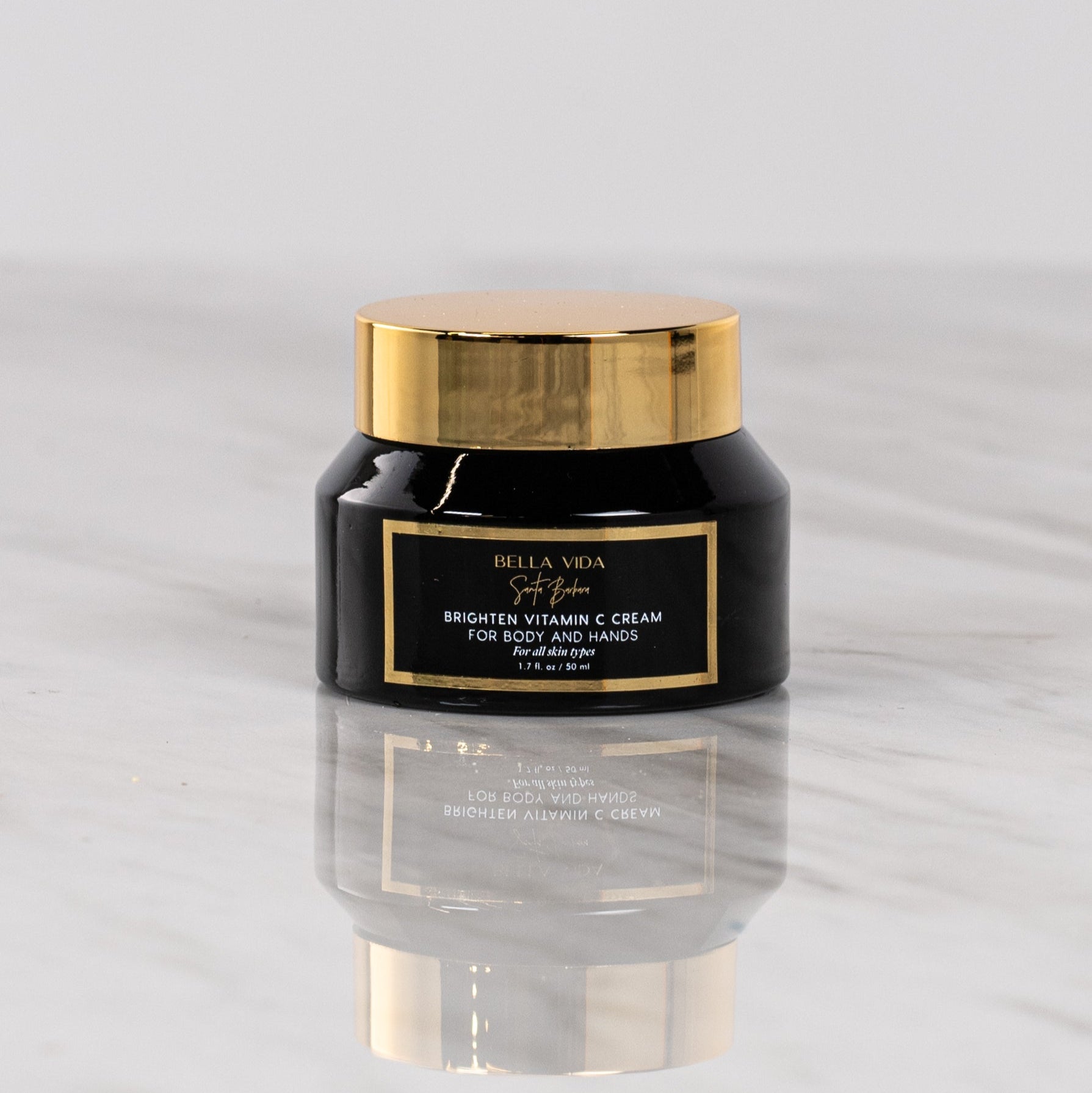 A jar of Bella Vida Santa Barbara Brighten Vitamin C Cream with a black label and gold lid, displayed on a marble surface.