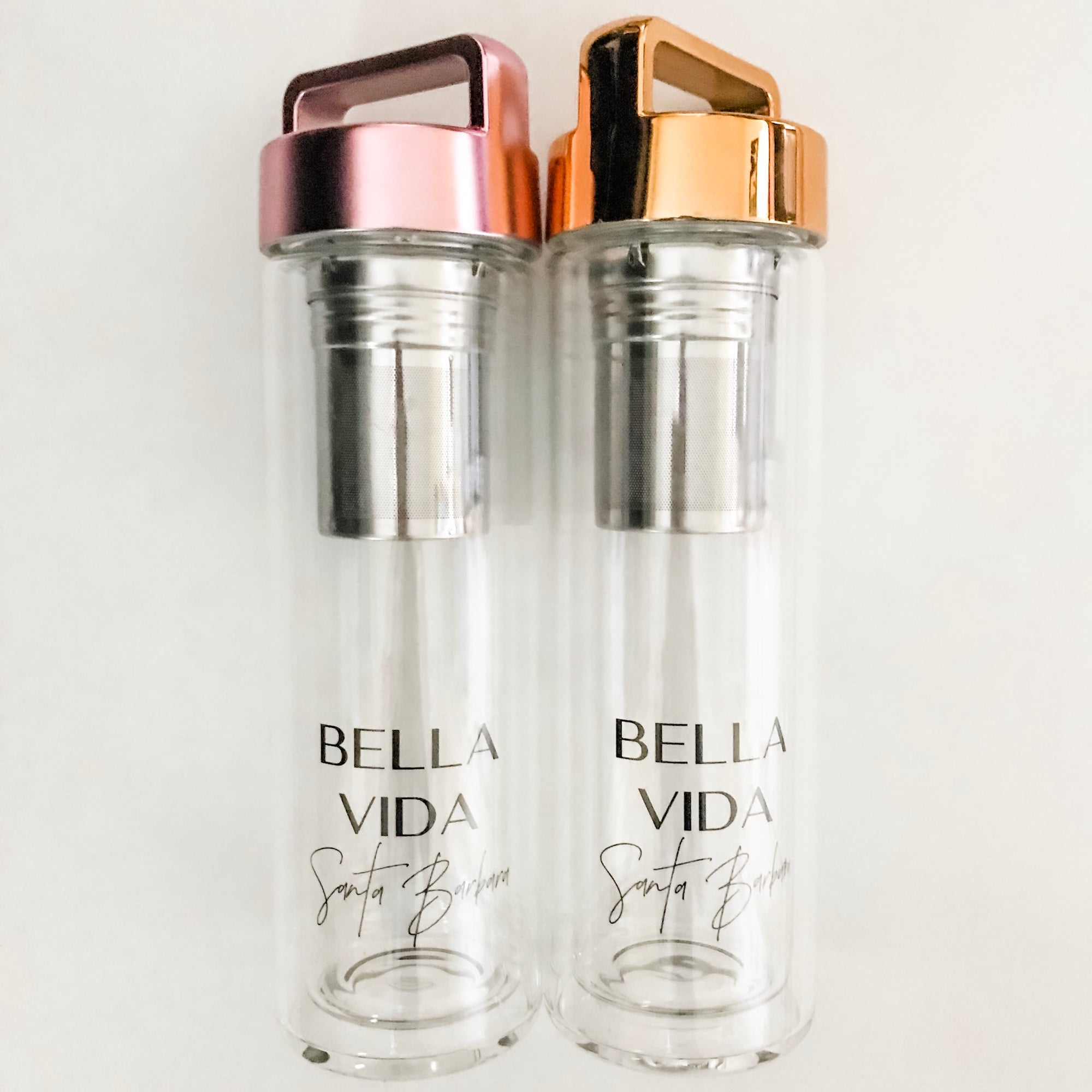 two glass water bottles by Bella Vida Santa Barbara one with gold lid one with pink lid