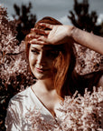 Woman with red hair shielding her eyes from the sun with a hand, surrounded by pink flowers.